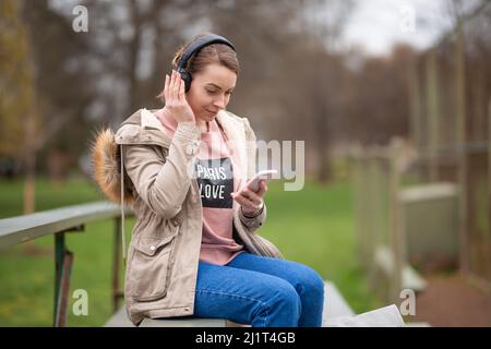 Fröhlich energisches Mädchen, auf einem Spaziergang im Park mit Kopfhörern und Telefon, hört Musik, mit leerem Raum, ein Spaziergang und Erholung im Freien, r Stockfoto