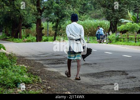 Ein alter indischer Mann, der lässige Tücher und eine Affenmütze trägt und einen Regenschirm hält, der alleine zu seinem Haus in Kolhapur, Indien, läuft Stockfoto