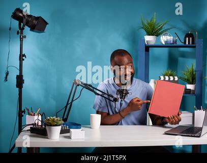 Porträt eines afroamerikanischen Vloggers, der eine Produktbewertung eines roten Buches vorführt, das mit dem Finger auf das Mockup-Cover im Vlogging-Studio zeigt. Influencer sitzt am Schreibtisch mit Podcast-Aufnahme-Setup und rezensieren Roman. Stockfoto