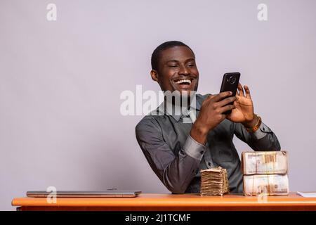 Ein junger, fröhlicher schwarzer Mann, der mit Geldpaketen im Büro sitzt, sein Telefon benutzt und ein Anlagekonzept entwickelt Stockfoto