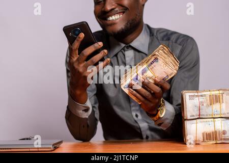 Ein junger, fröhlicher schwarzer Mann, der mit Geldpaketen im Büro sitzt, sein Telefon benutzt und ein Anlagekonzept entwickelt Stockfoto