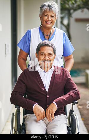 Mit einem Lächeln Hilfe leisten. Porträt eines älteren Mannes in einem Rollstuhl, der von einer Krankenschwester in einem Altersheim betreut wird. Stockfoto