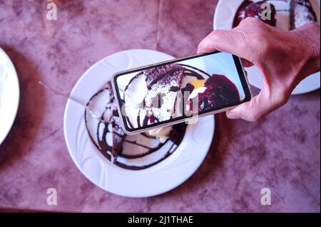 Hand des Mannes hält Handy und schießt Schokoladenkuchen Stockfoto