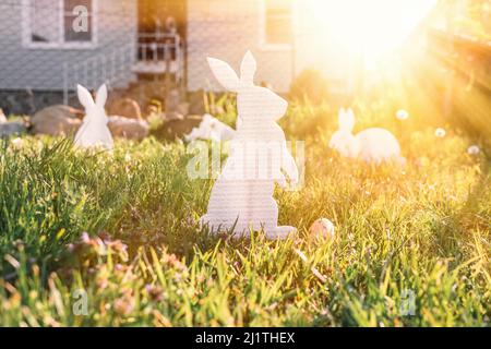 Das Konzept der Osterferien. Dekoratives Kunsthandwerk handgefertigt in Form von verschiedenen weißen Kaninchen, neben den bemalten Eiern im Gras. Stockfoto