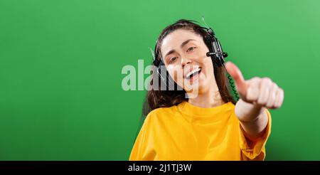 Online-Kurse und Lernen. Porträt einer jungen lächelnden Frau, die Kopfhörer trägt und den Daumen nach oben zeigt. Grüner Hintergrund. Banner. Das Konzept des Lernens Stockfoto