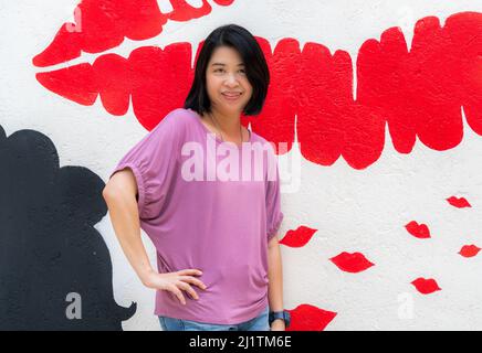 Porträt Asiatische Frau mittleren Alters mit schönen grafischen Wand in der Öffentlichkeit, lächelnd und posiert mit einer Hand auf der Taille, schwarze kurze Haare, trägt lässig, out Stockfoto