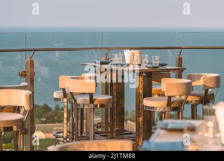 Stilvoller Tisch und Hocker eines Restaurants im Freien auf dem Dach eines Gebäudes, Geschirr auf dem Tisch, Glaszaun, verschwommener Hintergrund des Himmels Stockfoto