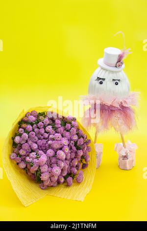 Bouquet von lila Chrysanthemen und Mädchen Ei in Rock und Hut auf gelbem Hintergrund. Ostern, Geburtstag, 8. März, Internationaler Frauentag, Muttertag Stockfoto