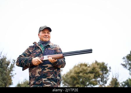 Männlicher Jäger in Camouflage-Oberbekleidung und Mütze, der eine Waffe trägt, während er im Herbstfeld herumhängt Stockfoto