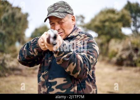 Älterer Mann in Tarnkleidung und Mütze, der an einem Herbsttag auf einem Feld auf die Kamera schiebt und mit der Schrotflinte auf die Kamera zeigt. Stockfoto