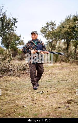 Ganzkörper gealterter männlicher Jäger in Camouflage-Oberbekleidung und Mütze, der Waffe trägt und wegschaut, während er Zeit in der herbstlichen Landschaft verbringt Stockfoto