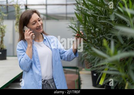 Eine junge Frau kauft in einem Blumengeschäft ein und wählt Zimmerpflanzen aus. Ruft mit seinem Smartphone an. Stockfoto