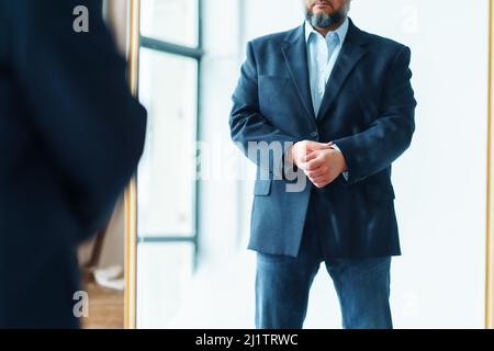 Beschnittenes Foto eines Geschäftsmannes mittleren Alters mit grauem Bart und blauer Anzugjacke, der drinnen vor dem Spiegel steht. Stockfoto