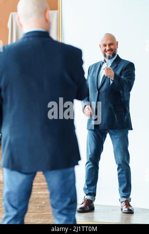 Porträt eines glücklichen Mannes mittleren Alters mit Glatze und grauem Bart, der eine blaue Jacke trägt und mit dem Finger auf die Spiegelreflexion zeigt. Stockfoto