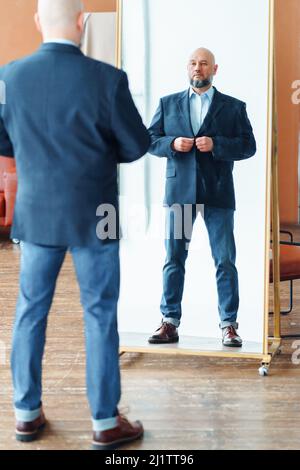 Porträt eines selbstbewussten Mannes mittleren Alters mit Glatze und grauem Bart, der eine blaue Jacke aufknöpft und Spiegelreflexion betrachtet. Stockfoto