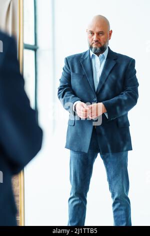 Porträt eines Mannes mittleren Alters mit Glatze und grauem Bart, der eine blaue Anzugjacke trägt und auf Spiegelreflexion grassiert. Stockfoto