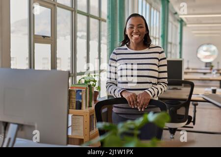 Lächelnde junge afrikanische Geschäftsfrau, die in einem Büro neben ihrem Schreibtisch steht Stockfoto