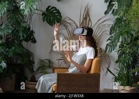 Junge, verblüffte Gärtnerin testet VR-Helm für den Geschäftsbetrieb, während sie im Gewächshaus sitzt Stockfoto