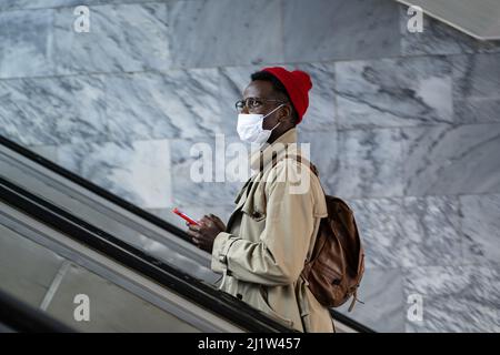 Nachdenklicher afroamerikanischer Tausendjähriger Mann mit Smartphone in der U-Bahn mit Rolltreppe und Schutzmaske Stockfoto
