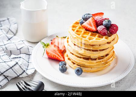Hausgemachte Waffeln mit Beeren, Puderzucker und Ahornsirup auf einem weißen Teller, grauer Hintergrund. Stockfoto