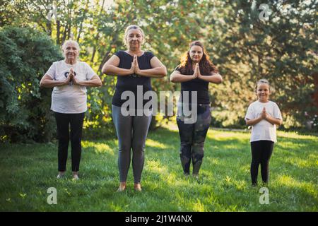 Wenige Generationen von Frauen aus einer Familie, die in Sukhasana zusammen stehen, posieren auf einem Hinterhof voller grünem Gras und Bäumen, die Yoga machen. Zeit verbringen Stockfoto