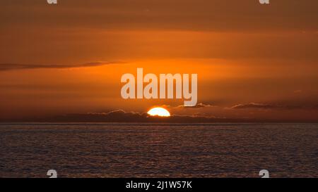 Blick auf den Sonnenuntergang mit Sonnenuntergang am Horizont zwischen niedrigen Wolken mit dramatisch orangefarbenem Himmel von der Küste von Taurito, Gran Canaria aus gesehen. Stockfoto