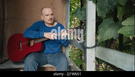 Der Mensch leitet eine Online-Akustikgitarre-Lektion von der Veranda seiner Vorstadtfarm aus Stockfoto