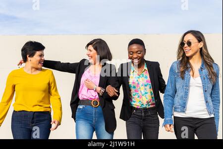 Gruppe fröhlicher multirassischer Freundinnen, die sich unterhalten und in der Nähe der weißen Mauer auf der sonnigen Straße der Stadt während des Treffens zusammen gehen Stockfoto