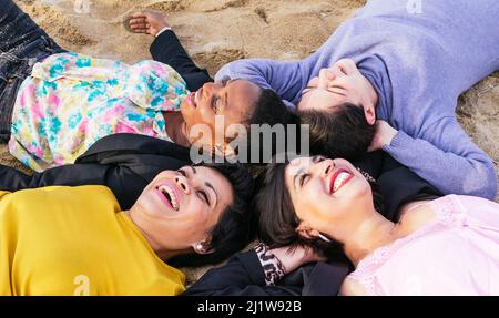 Draufsicht Gruppe positiver multirassischer Freundinnen, die am Sommertag auf die Kamera blicken, während sie zusammen am Sandstrand liegen Stockfoto