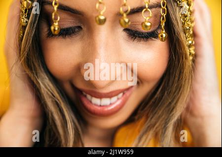 Crop optimistische hispanische Bauchtänzerin mit stilvollem Make-up und Kette-Stirnband-Mütze, die im Studio vor hellgelbem Hintergrund fröhlich lächelt Stockfoto