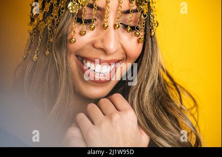 Crop optimistische hispanische Bauchtänzerin mit stilvollem Make-up und Kette-Stirnband-Mütze, die im Studio vor hellgelbem Hintergrund fröhlich lächelt Stockfoto