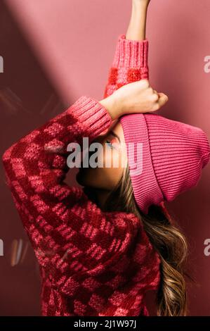 Seitenansicht eines attraktiven Weibchens mit Make-up in farbenprächtiger rosa Mütze und Strickjacke auf rosa Hintergrund mit erhobenem Arm in hellem Studio Stockfoto