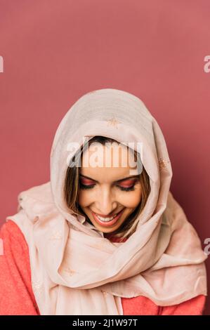 Friedliche muslimische Frau mit Make-up-Abdeckung Gesicht mit langen hellen Kopftuch, während auf rosa Hintergrund Blick auf die Kamera im Studio stehen Stockfoto