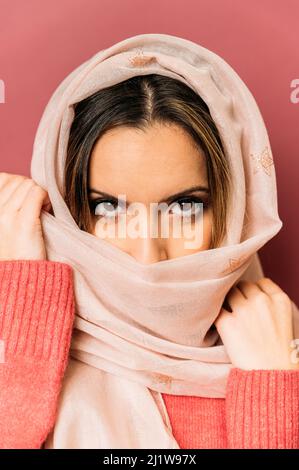 Friedliche muslimische Frau mit Make-up-Abdeckung Gesicht mit langen hellen Kopftuch, während auf rosa Hintergrund Blick auf die Kamera im Studio stehen Stockfoto