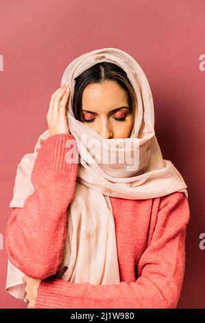 Friedliche arabische Frau mit Make-up bedeckendem Gesicht mit langem leichten Kopftuch, während sie auf rosa Hintergrund mit geschlossenen Augen im Studio steht Stockfoto