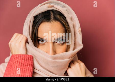Friedliche muslimische Frau mit Make-up-Abdeckung Gesicht mit langen hellen Kopftuch, während auf rosa Hintergrund Blick auf die Kamera im Studio stehen Stockfoto