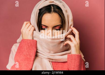 Friedliche arabische Frau mit Make-up bedeckendem Gesicht mit langem leichten Kopftuch, während sie auf rosa Hintergrund mit geschlossenen Augen im Studio steht Stockfoto