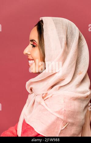 Seitenansicht einer optimistischen Frau mit farbenfrohem Make-up und leichtem Kopftuch, während sie im Studio auf rosa Hintergrund steht Stockfoto