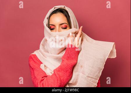 Friedliche arabische Frau mit Make-up bedeckendem Gesicht mit langem leichten Kopftuch, während sie auf rosa Hintergrund mit geschlossenen Augen im Studio steht Stockfoto