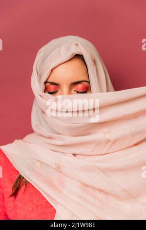 Friedliche arabische Frau mit Make-up bedeckendem Gesicht mit langem leichten Kopftuch, während sie auf rosa Hintergrund mit geschlossenen Augen im Studio steht Stockfoto