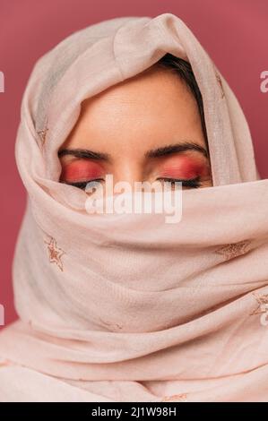 Friedliche arabische Frau mit Make-up bedeckendem Gesicht mit langem leichten Kopftuch, während sie auf rosa Hintergrund mit geschlossenen Augen im Studio steht Stockfoto