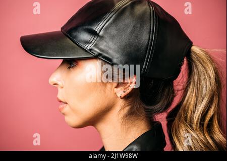 Seitenansicht des langhaarigen blonden hispanischen weiblichen Models mit dunkler Lederjacke und Mütze auf rosa Hintergrund im Studio Stockfoto