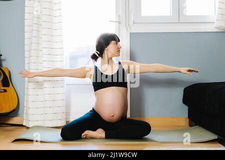 Friedliche Schwangeren im aktiven Ohr sitzen in Yoga-Pose mit ausgestreckten Armen während der Sitzung im Wohnzimmer auf einer Matte Stockfoto