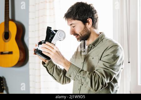 Aufmerksamer junger ethnischer Mann mit dunklem Haar und Bart in legerer Kleidung, der VR-Brille untersucht, während er bei Tageslicht zu Hause steht Stockfoto
