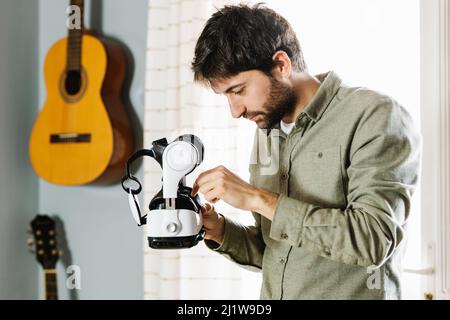 Aufmerksamer junger ethnischer Mann mit dunklem Haar und Bart in legerer Kleidung, der VR-Brille untersucht, während er bei Tageslicht zu Hause steht Stockfoto