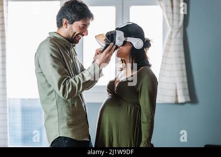 Seitenansicht eines fröhlichen jungen hispanischen Mannes, der hell lächelt, während er die VR-Brille vom Kopf einer positiven Schwangeren auszieht, nachdem er die virtuelle Realität erkundet hat Stockfoto