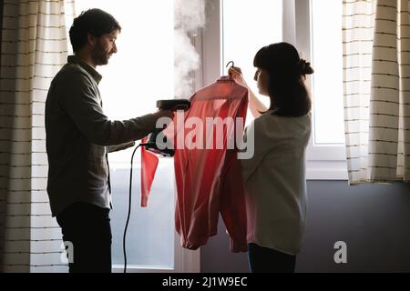 Seitenansicht einer Schwangeren, die in der Nähe eines Mannes mit Bügelhemd und manuellem Dampfgarer im Haus steht Stockfoto