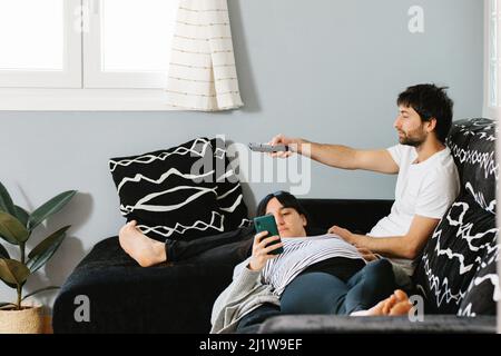 Ganzer Körper einer jungen, ethnischen, schwanger barfüßigen Dame, die Mobiltelefon benutzt, während sie auf den Knien des Mannes auf dem Sofa sitzt und die Fernsehkanäle wechselt Stockfoto