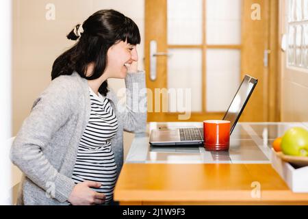 Seitenansicht einer positiven jungen, hispanischen, Schwangeren Freiberuflerin mit dunklem Haar und lächelndem Bauch, während sie an einem Laptop am Tisch arbeitete Stockfoto