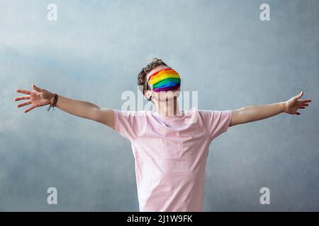 Anonymes Transgender-Gesicht mit farbenfroher Schutzmaske mit LGBT-Regenbogenmuster, während es mit ausgebreiteten Armen auf grauem Hintergrund steht Stockfoto